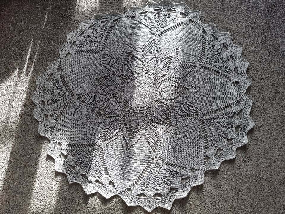 Overhead view of a large round grey crochet doily with intricate lace floral patterns on a carpet with sunlight.