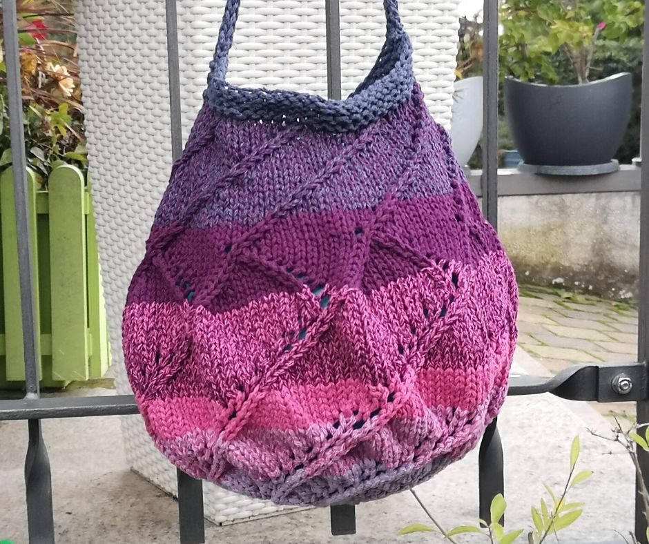 Gestrickte Ombre-Tasche in Blau, Violett und Pink mit Zopf- und Lochmustern, hängend an einem Metallgeländer.