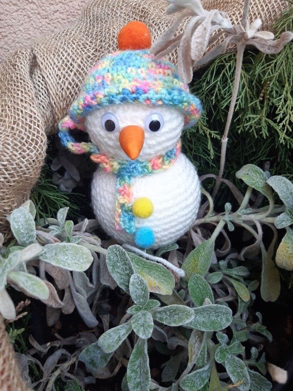 Gehäkelter weißer Schneemann mit bunter Mütze, Schal und orangefarbener Nase zwischen frostigen Pflanzen