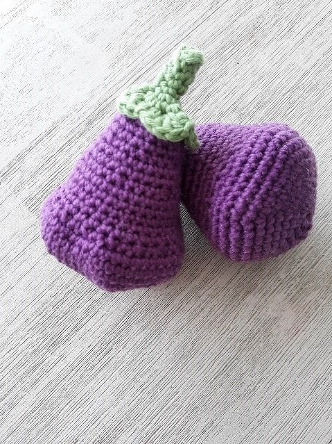 Zwei lila Amigurumi-Auberginen mit grünem Blattstiel nebeneinander auf hellem Untergrund