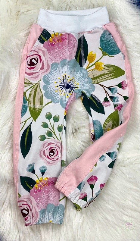 Bequeme Babyhose mit großem Blumenmuster, rosa Seitenstreifen und weißem Bund, liegend auf flauschigem Untergrund.
