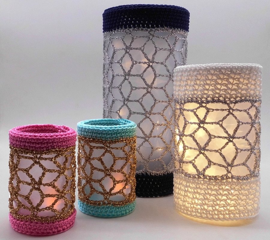 Vier gehäkelte Windlichter in verschiedenen Größen auf hellem Hintergrund, innen beleuchtet. Farben: Rosa, Türkis, Dunkelblau und Weiß mit glänzenden Garnakzenten.