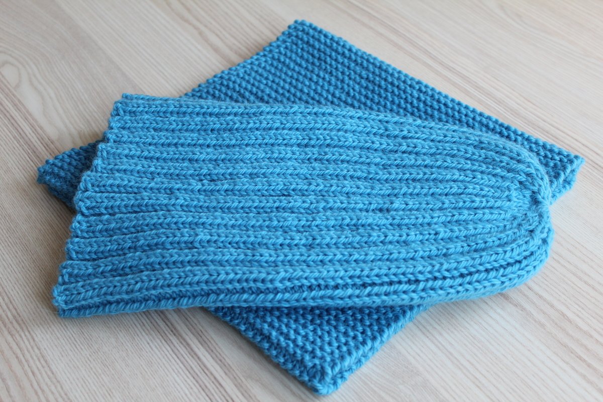 Nahaufnahme: Rippenmuster einer blauen Beanie und kraus-rechts gestricktes Tuch auf hellem Holz