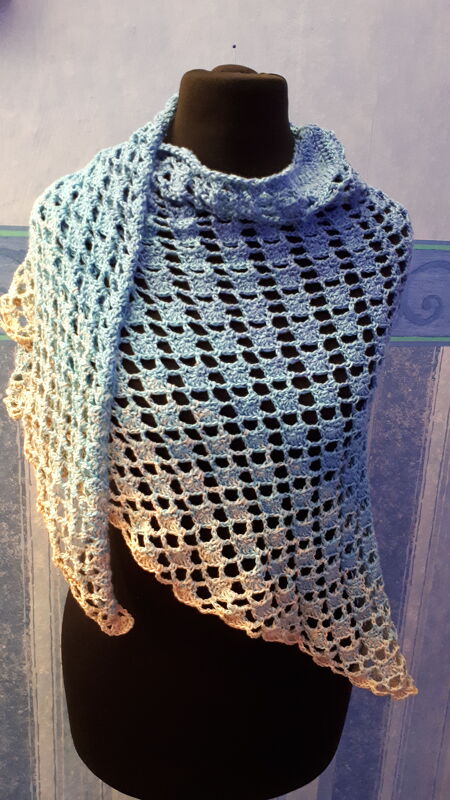 Dreieckiges Häkeltuch mit blau-beigem Farbverlauf und Lochmuster auf einer schwarzen Schaufensterpuppe drapiert.