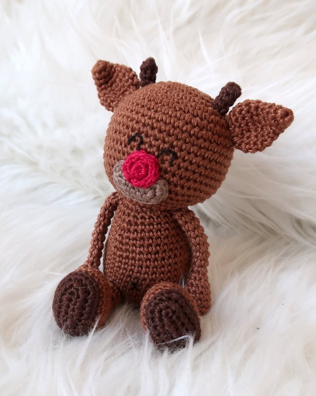 Gehäkeltes braunes Rentier-Amigurumi mit roter Nase auf weißem, flauschigem Untergrund