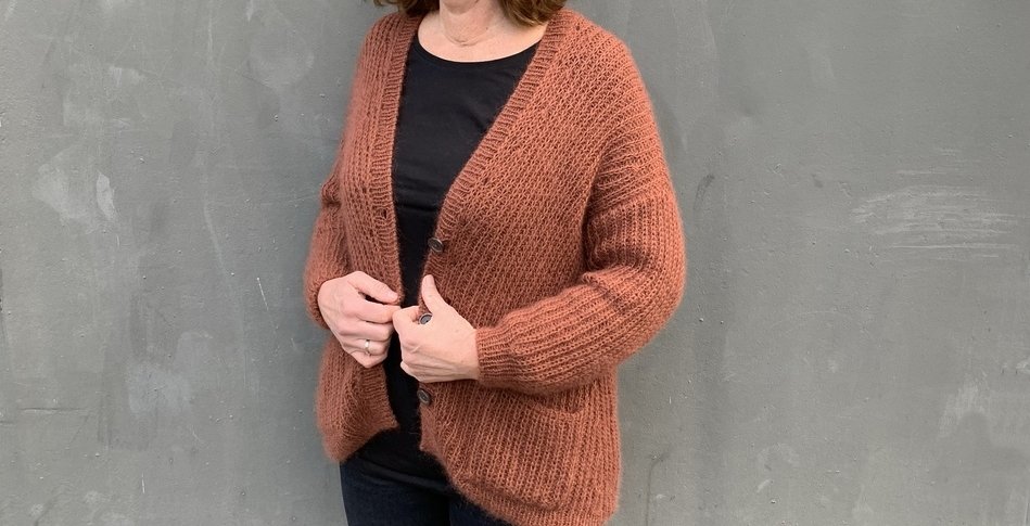 Person trägt eine rostbraune, locker gestrickte Strickjacke mit Knopfleiste und aufgesetzten Taschen vor einer grauen Wand