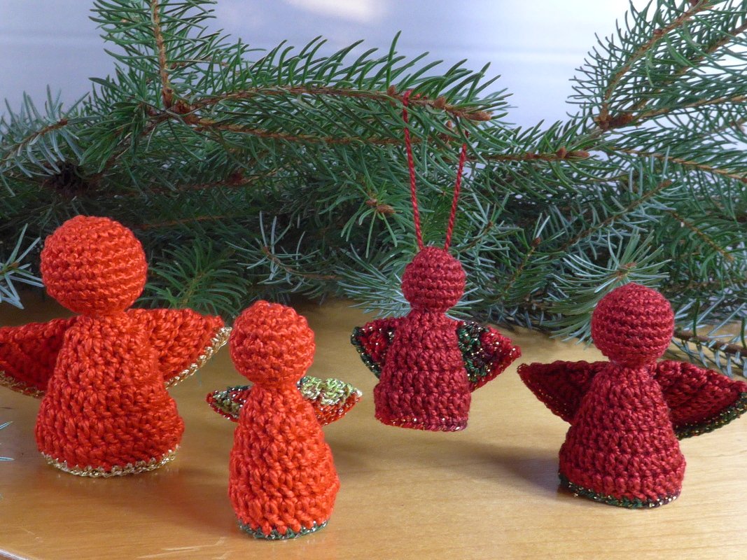 Vier gehäkelte rote Weihnachtsengel, drei stehend und einer hängend, vor Tannenzweigen auf einer Holzfläche.