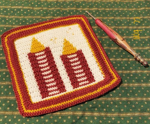 Häkelquadrat mit zwei stilisierten Kerzen in Rot und Gelb auf grün gemustertem Stoff mit Häkelnadel