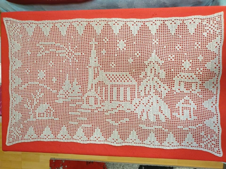 Weißer rechterckiger Tischläufer im Filet-Häkelmuster mit Dorfmotiv auf rotem Stoff.