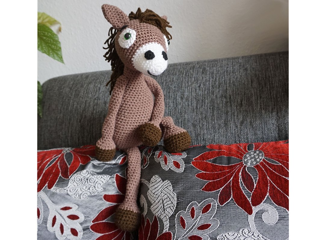 Brown crocheted amigurumi horse with dark mane and hooves, white muzzle, green eyes, on a red and grey floral cushion.