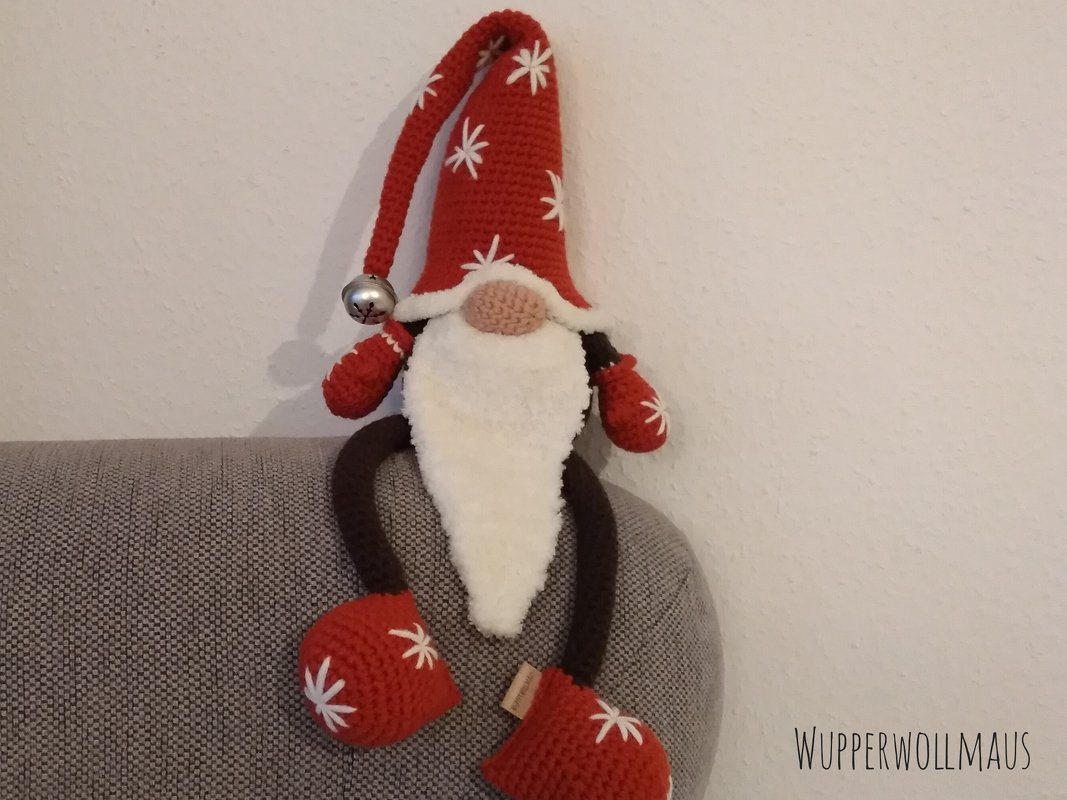 Gehäkelter Weihnachtswichtel mit roter Zipfelmütze, weißem Bart und Glocke, sitzend auf einer Sofalehne