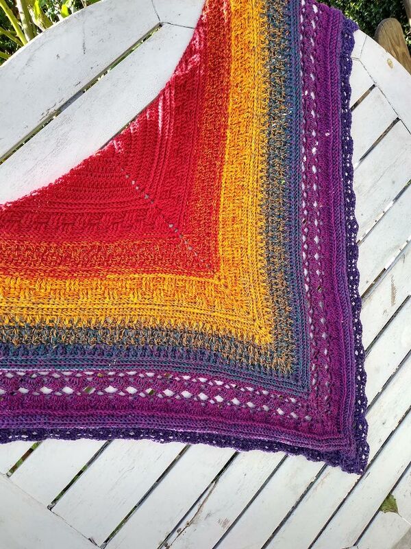 Gehäkeltes Dreieckstuch mit Farbverlauf von Rot zu Violett, Strukturmaschen, Lochreihe und gezackter Kante auf weißem Holz