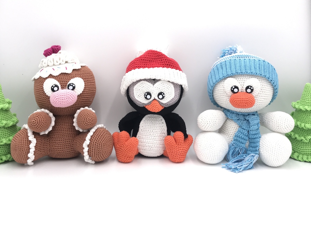 Three crocheted Christmas amigurumi figures: gingerbread man, penguin with Santa hat, and snowman with blue hat and scarf, with small green crochet trees at each side.