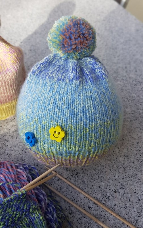 Gestrickte Kindermütze mit blau-gelbem Garn, mehrfarbigem Bommel und zwei Blumenknöpfen; Stricknadeln und Wolle daneben