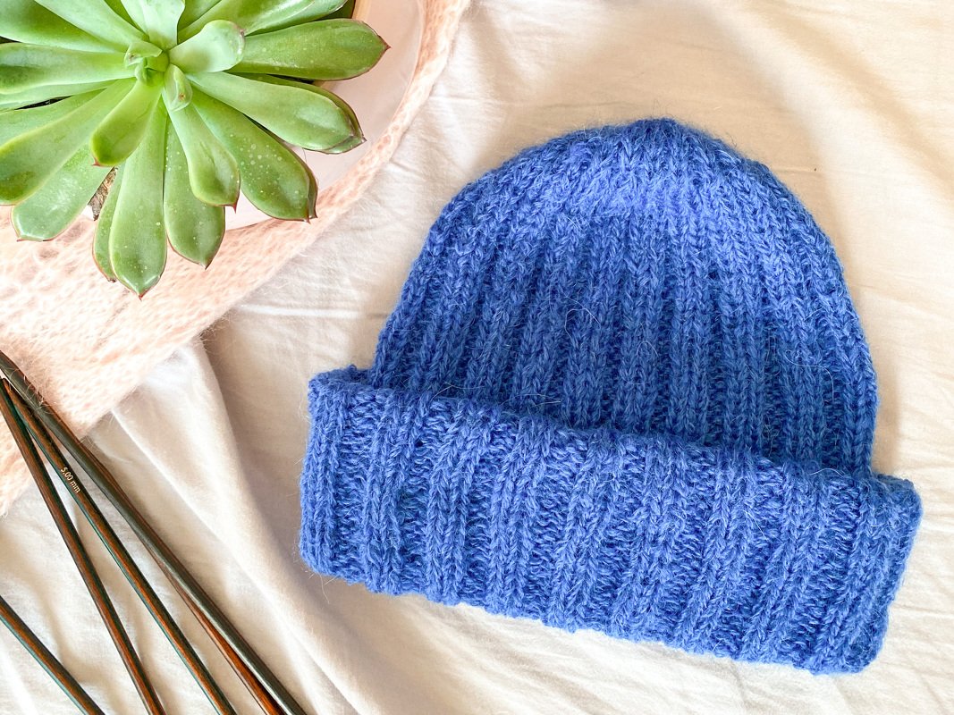 Nahaufnahme einer blau gestrickten Rippmütze mit umgeschlagenem Bund, Stricknadeln liegen daneben