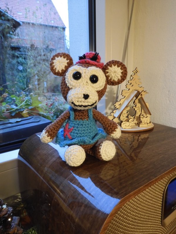 Gehäkelter braun-cremefarbener Amigurumi-Affe mit blauem Kleid und roter Mütze, sitzend auf Holz vor Fenster