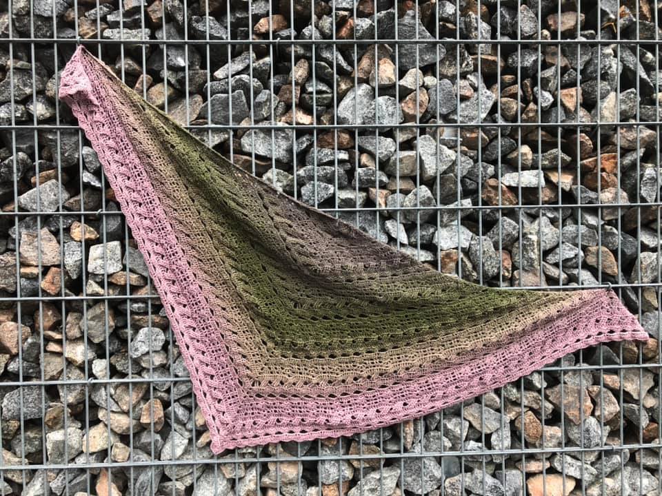 Gehäkeltes Dreieckstuch mit Farbverlauf in Rosa, Grün und Braun, liegend vor einer Gabionenwand