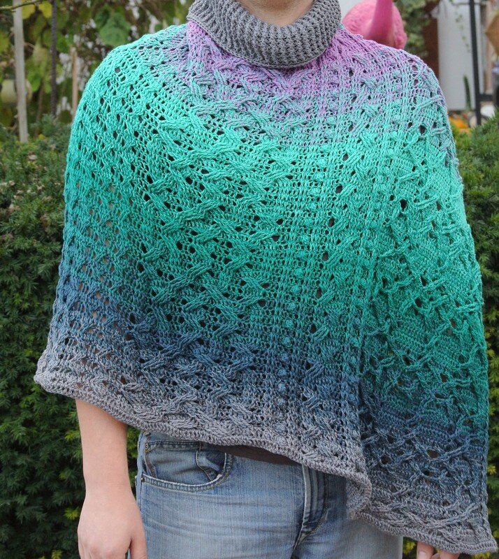 Person trägt gehäkelten Poncho mit Farbverlauf von Grau zu Türkis und Violett sowie grauem Rollkragen.
