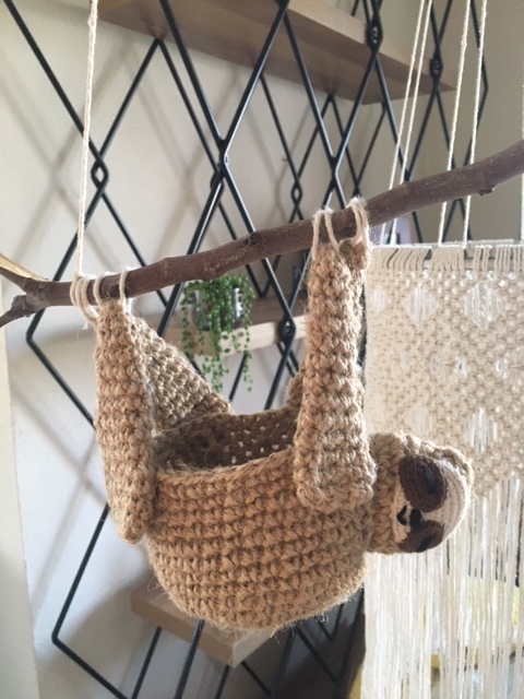 Tan crocheted sloth hanging basket with limbs looped over a wooden branch