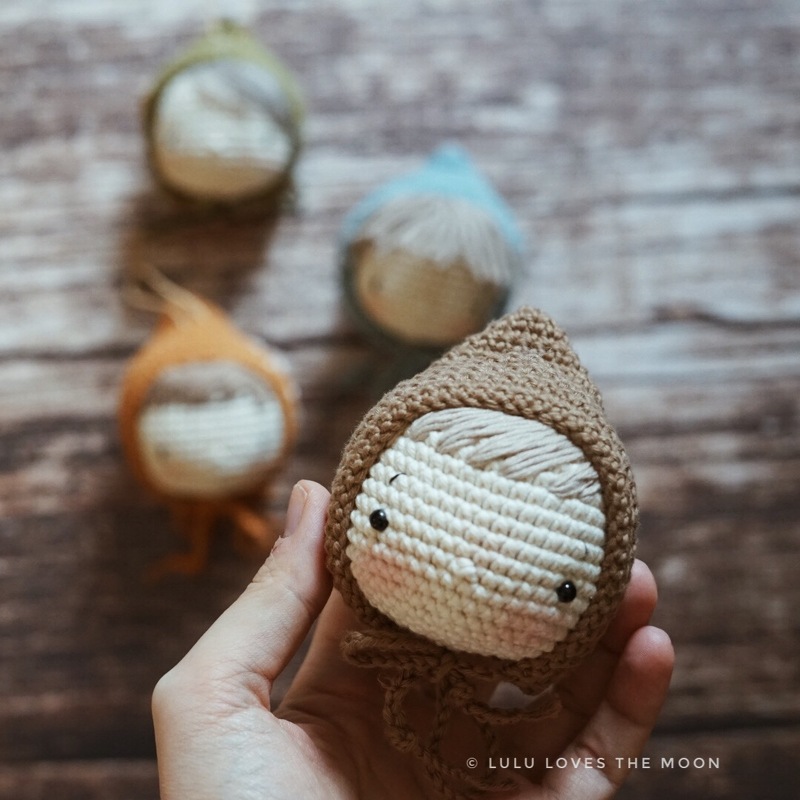 A brown crocheted gnome-like figure with an acorn hood held in a hand, with other colorful crochet figures blurred in the background.