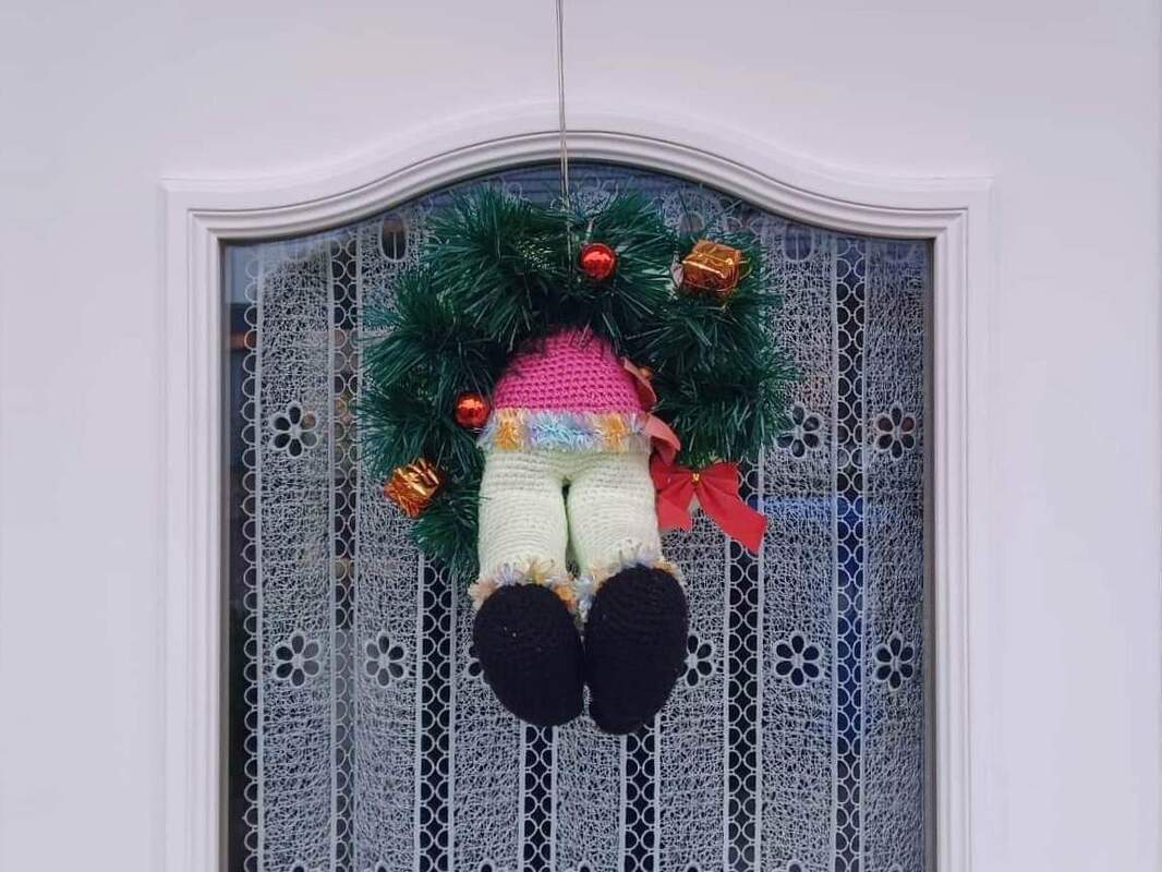 Gehäkelter grüner Türkranz mit hängenden Puppenbeinen, roten Kugeln und roter Schleife vor Spitzenvorhang