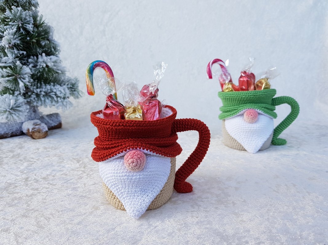 Gehäkelter Wichtelbecher in Rot mit Bonbons, im Hintergrund grüner Wichtelbecher, auf hellem Untergrund neben verschneigtem Tannenzweig