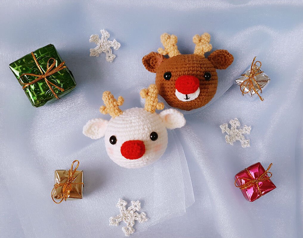 Two crocheted amigurumi reindeer head ornaments, one white and one brown, with red noses and small antlers, surrounded by tiny wrapped gifts and crochet snowflakes on pale blue fabric.