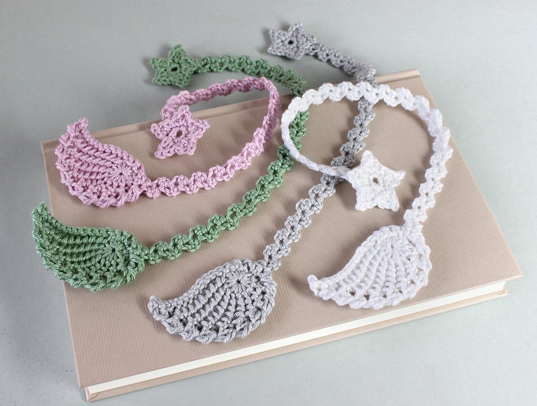 Vier gehäkelte Lesezeichen in Weiß, Hellrosa, Salbeigrün und Hellgrau mit Flügel- und Sternmotiven auf einem Buch.