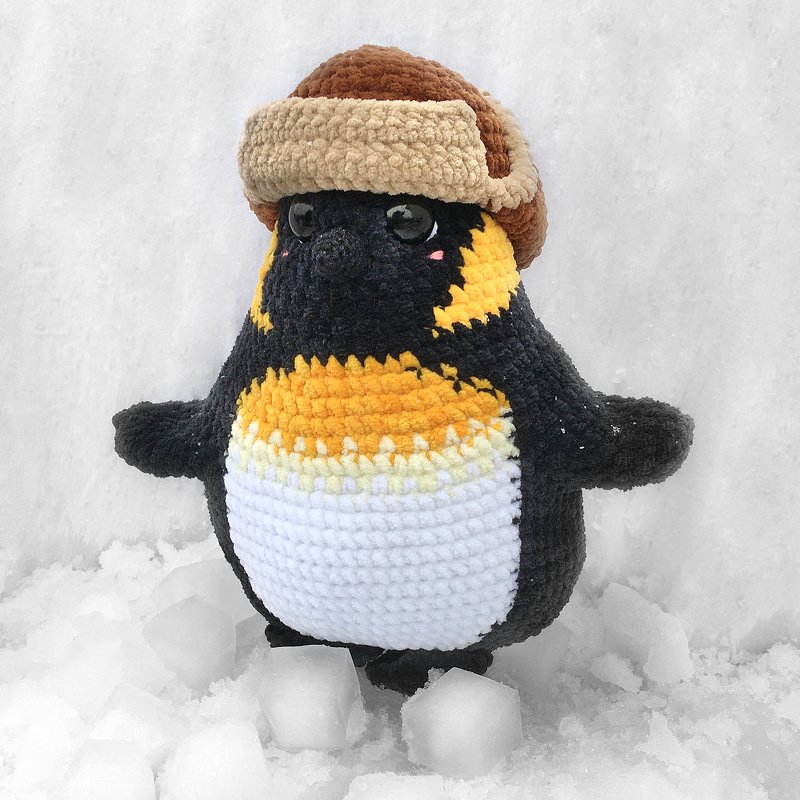 Nahaufnahme eines gehäkelten Pinguins mit Ohrenklappenmütze und rosa Wangen im Schnee