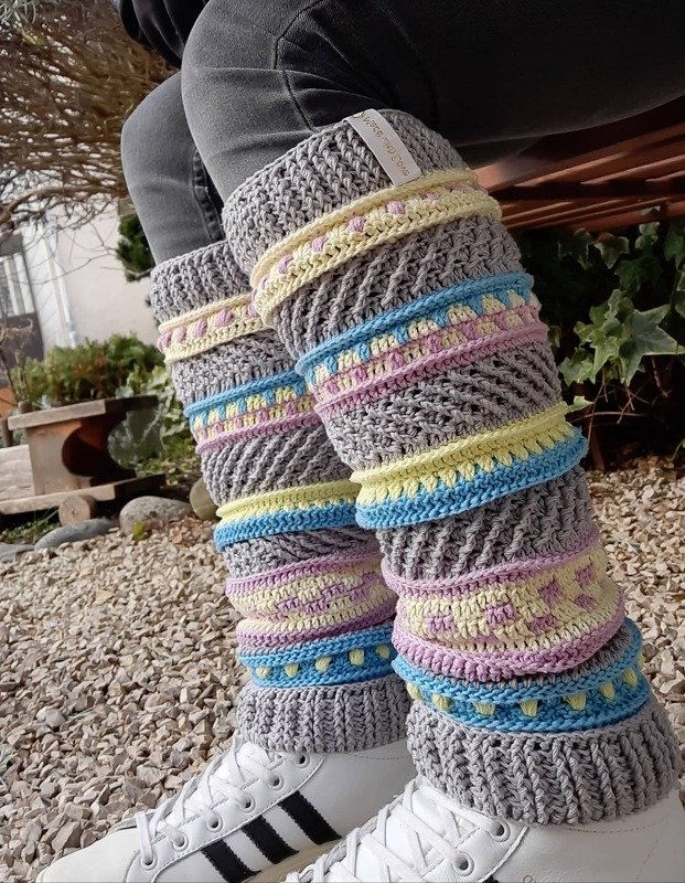 Gehäkelte graue Beinstulpen mit pastellgelben, hellblauen und rosafarbenen Streifen über dunklen Hosen und weißen Sneakern
