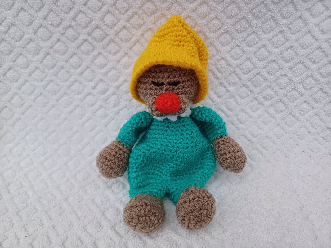 Gehäkelte Kuschelpuppe in Türkis mit gelber Zipfelmütze und roter Nase auf weißer, strukturierter Unterlage.