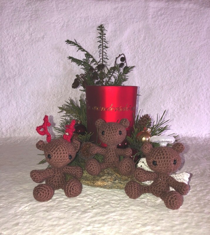 Drei braune gehäkelte Amigurumi-Bären (Rentier, Engel, klassischer Bär) vor rotem Kerzenhalter und Tannengrün