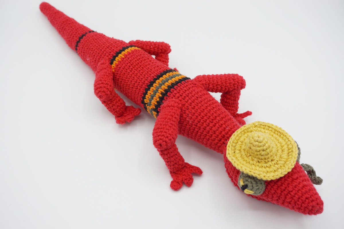 Red crocheted amigurumi lizard with multicolor stripes wearing a small yellow sombrero on a white background.