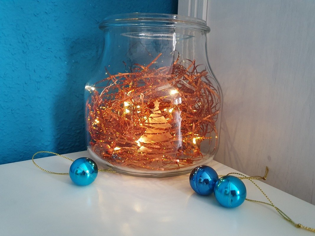 Transparentes Glasgefäß mit kupferfarbenem Lametta, Lichterkette und ausgeschnittener Tannenbaum-Silhouette, davor drei türkisblaue Weihnachtskugeln.