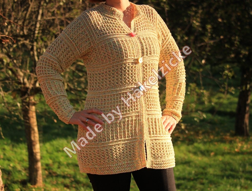 Beigefarbene lange gehäkelte Damenjacke mit verschiedenen Häkelstrukturen und Knöpfen, draußen fotografiert.