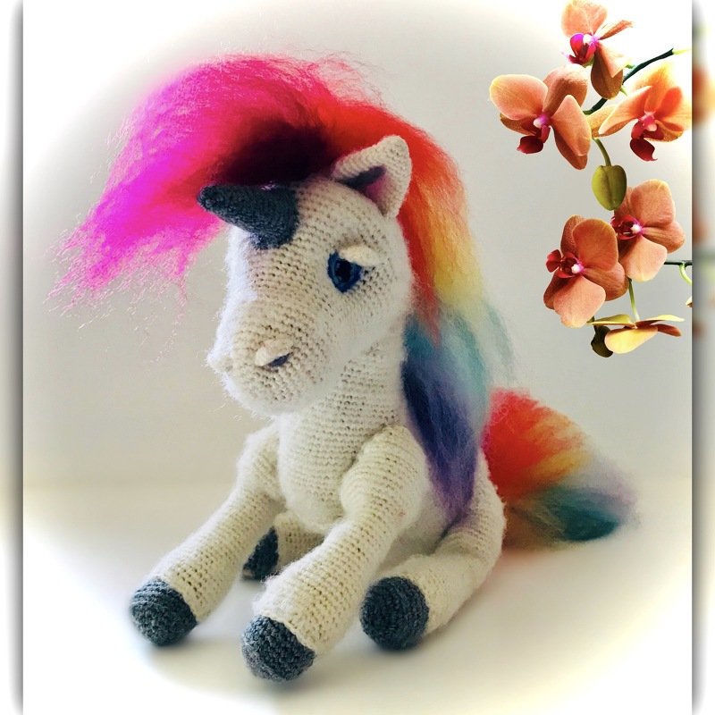 Weißes gehäkeltes Einhorn mit Regenbogenmähne, grauem Horn und blauen Augen, daneben orange Orchideen.