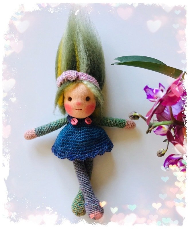 Gehäkelte Amigurumi-Puppe mit grünem Haar, blauem Kleid und rosa Stirnband neben violetten Orchideen