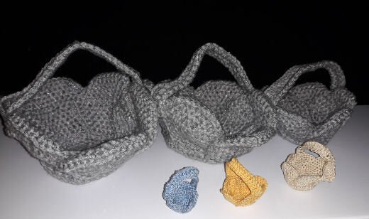 Gehäkelte Körbe in Grau mit Griffen, davor drei kleine gehäkelte Utensilos in verschiedenen Farben