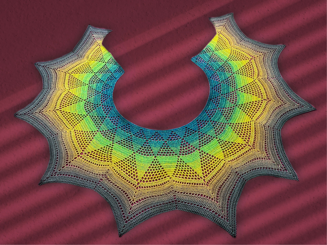 Lacy crocheted shawl with gradient colors from charcoal to yellow, green, and teal, featuring a starburst scalloped edge on a maroon background.
