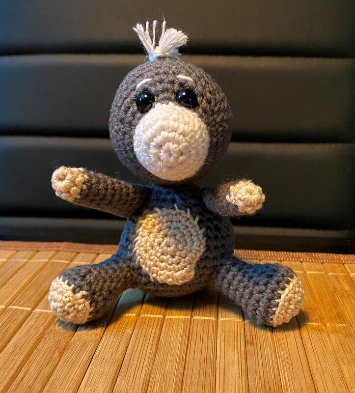 Gehäkeltes graues Amigurumi-Tier mit weißer Schnauze, dunklen Augen und kleinem Haarbüschel sitzend auf einer Bambusmatte.
