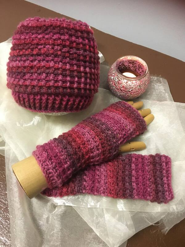 Gehäkelte Mütze und fingerlose Handschuhe (Stulpen) in Rosa- und Beerentönen, auf Tisch neben Kerze präsentiert
