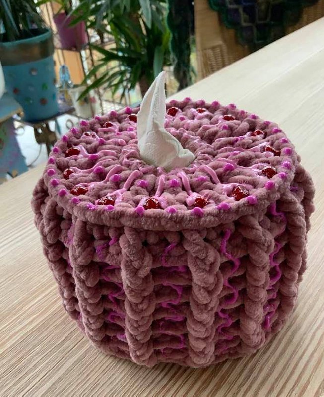 Runder gehäkelter Bezug für eine Taschentuchbox in Rosa mit roten Verzierungen und einem weißen Taschentuch, das oben herausragt.