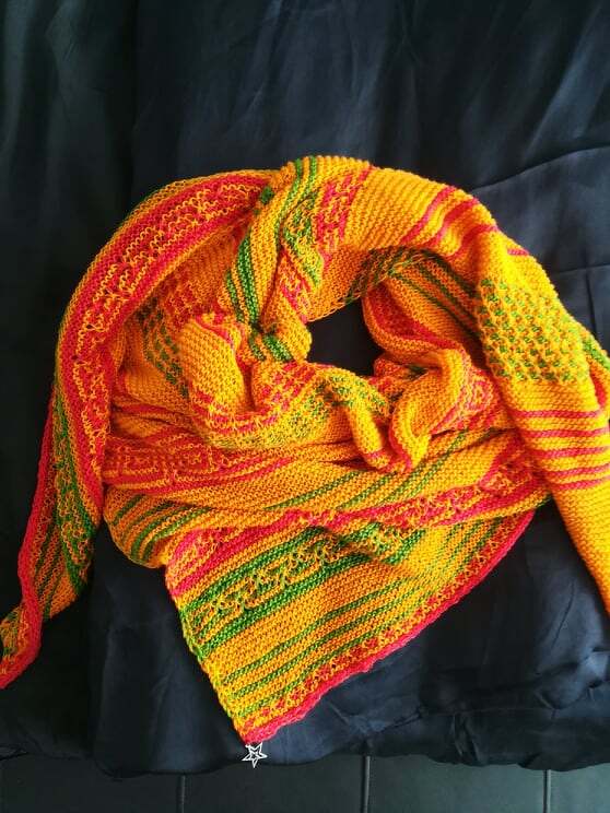 Gestricktes Dreieckstuch in Gelb, Orange und Grün mit Streifen und kleinen Lochmustern, zusammengerollt auf dunklem Stoff