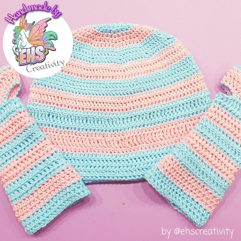 Gehäkelte Kindermütze und zwei Armstulpen in rosa- und hellblauen Streifen auf rosafarbenem Hintergrund.