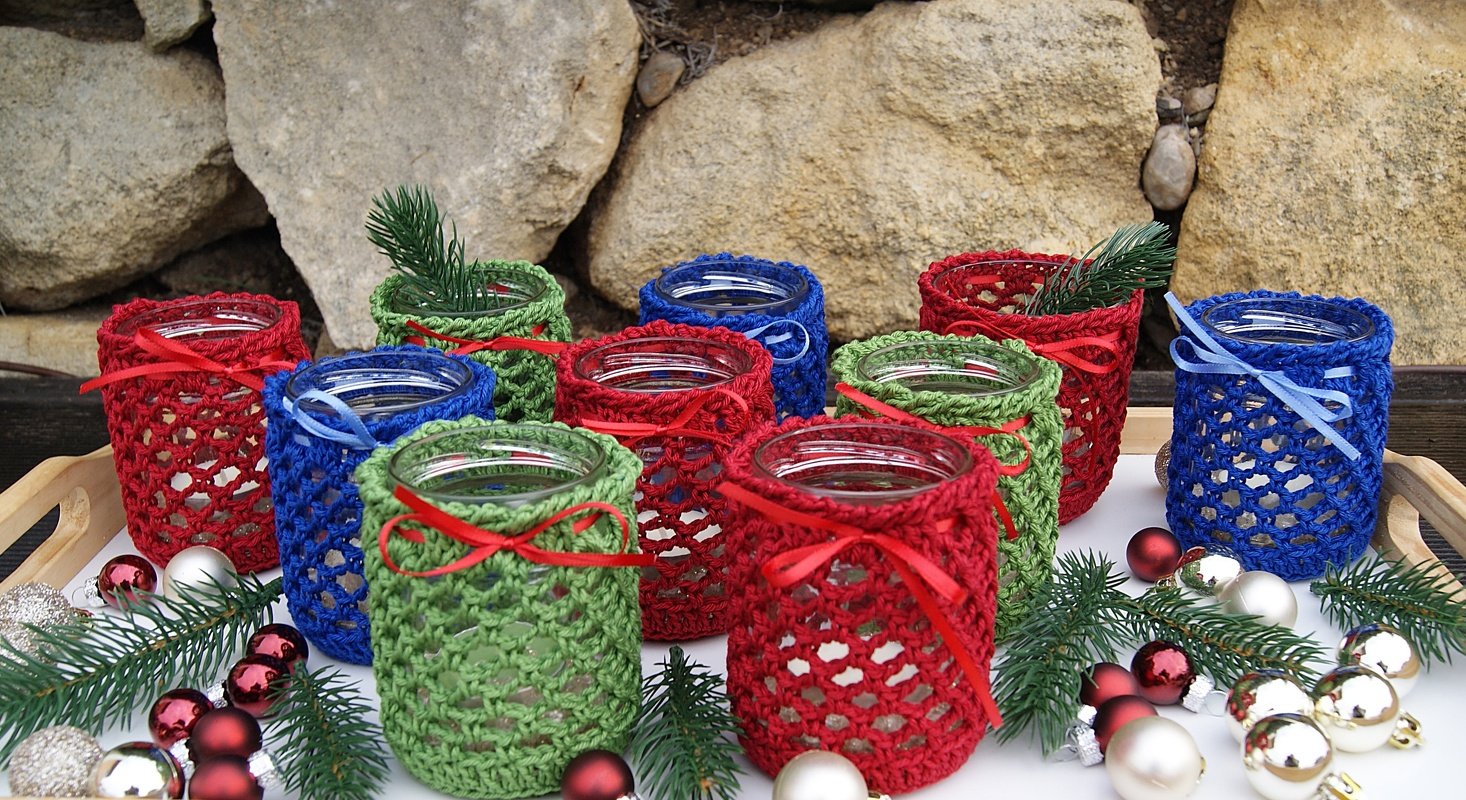 Glasgefäße mit gehäkelten Bezügen in Rot, Blau und Grün, dekoriert mit Schleifen, Tannenzweigen und Kugeln auf einem Tablett vor einer Steinwand.