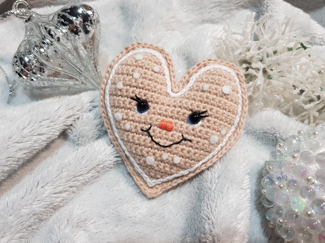 Gehäkeltes Herz in Lebkuchenoptik mit Gesicht auf Plüschdecke neben Weihnachtskugel