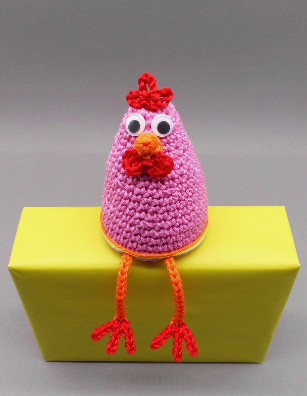 Rosa gehäkeltes Huhn mit Wackelaugen und orangen Beinen auf gelbem Kasten