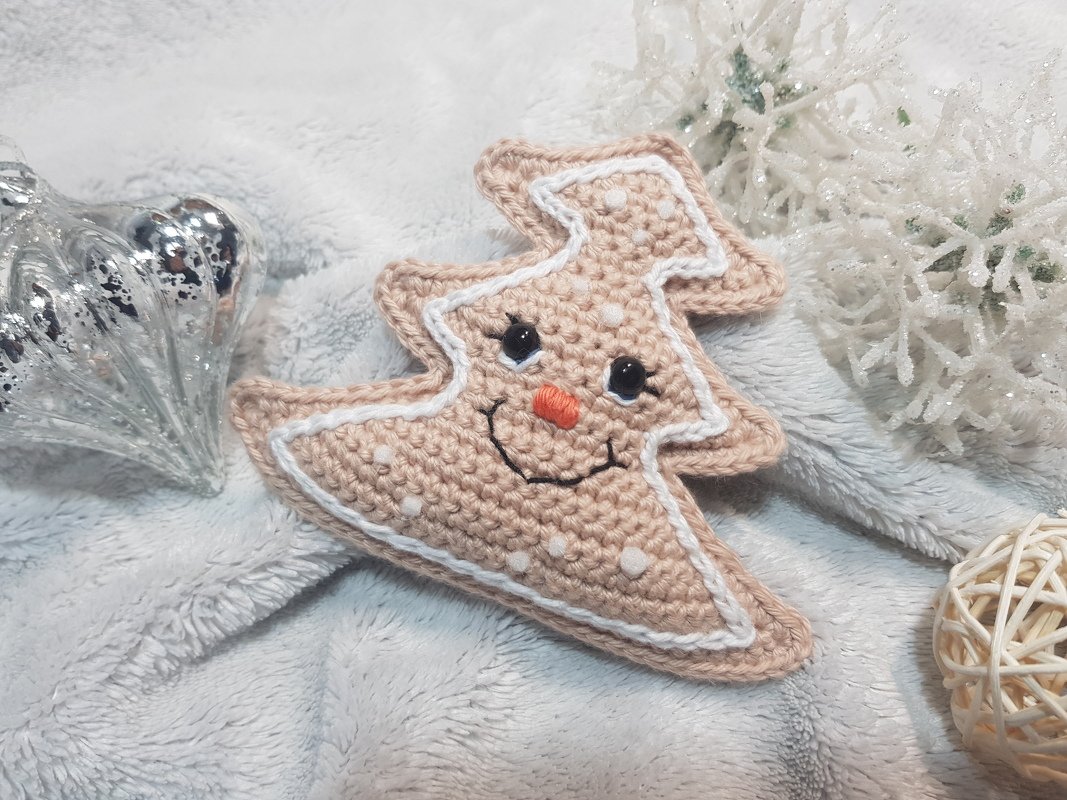 Beiger gehäkelter Weihnachtsbaum-Anhänger mit weißer Umrandung und lächelndem Gesicht auf flauschigem Stoff neben einer silbernen Kugel