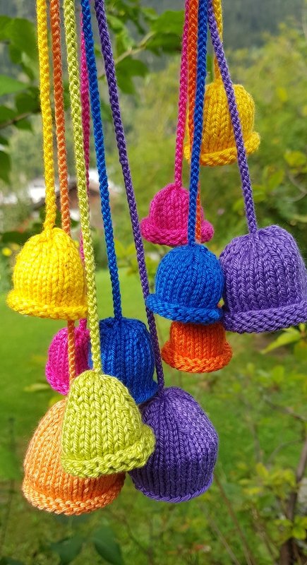 Mehrere bunte gestrickte Glöckchen hängen an farbigen Schnüren vor grünem Hintergrund.