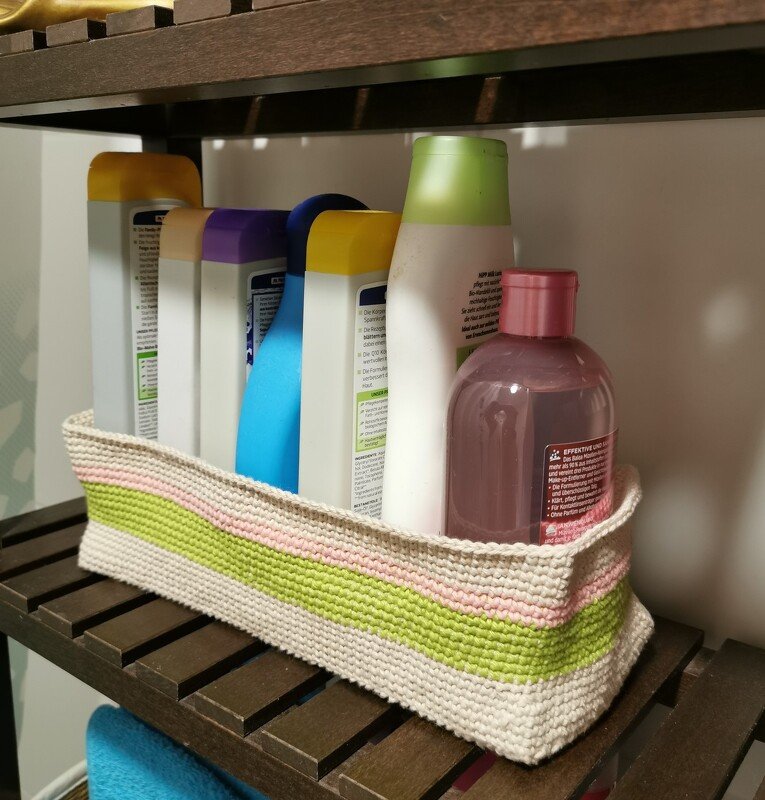 Gehäkelter Korb in Beige mit grünen und rosa Streifen, gefüllt mit Badezimmerflaschen auf einem Holzregal